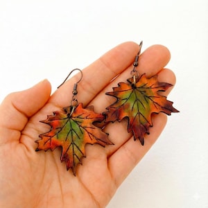 May include: A pair of leaf-shaped earrings. The earrings are in the shape of maple leaves, with a color gradient of green, orange, and brown. The earrings are held by a person's hand.