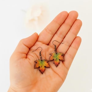 May include: A pair of maple leaf earrings. The earrings feature a detailed design with a gradient of colors, including green, yellow, orange, and red, and are suspended from bronze-colored hooks. The earrings are held in a hand against a white background.
