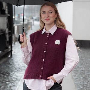 Op de afbeelding: Een bordeauxrode gebreide vest met een wit label wordt gedragen over een roze gestreept button-down overhemd. Het vest heeft witte knopen. De persoon houdt een zwarte paraplu vast.