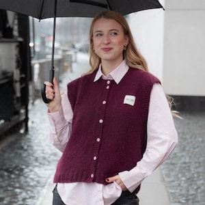 Könnte beinhalten: Eine weinrote Strickweste mit weißem Etikett, getragen über einem rosa gestreiften Hemd. Die Weste hat weiße Knöpfe und einen runden Ausschnitt. Die Person hält einen schwarzen Regenschirm.