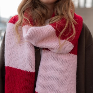 Könnte beinhalten: Ein grob gestrickter Schal mit abwechselnden roten und hellrosa Streifen. Der Schal ist um den Hals gewickelt und zeigt das auffällige Colorblocking. Das dicke Garn erzeugt einen gemütlichen, strukturierten Look, ideal für kaltes Wetter.