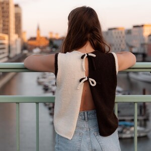 Könnte beinhalten: Ein ärmelloses, zweifarbiges Strickoberteil in Creme und Dunkelbraun mit Schleifendetail am Rücken. Das Oberteil wird mit hellblauen Jeans kombiniert. Das Bild wurde im Freien mit einer Stadtkulisse aufgenommen.