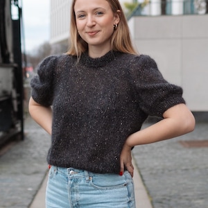 May include: A dark gray sweater with short, puffed sleeves and a speckled pattern. The sweater is paired with light blue denim jeans. The woman is smiling and standing outside.