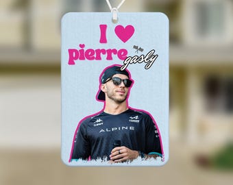 May include: Rectangular car air freshener with the text "I love Pierre Gasly" in pink and white. The air freshener features a photo of Pierre Gasly wearing a black cap and sunglasses. The background is a light blue.