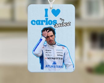 May include: Rectangular car air freshener with the text "I love Carlos Sainz" in blue. The air freshener features a photo of a man in a white and blue racing suit. The background is a blurred image of a house and a car's rearview mirror.