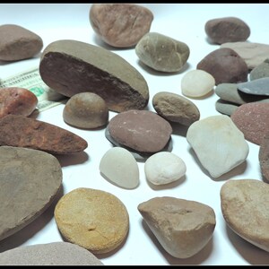 Pu&ograve; includere: Una collezione di pietre lisce e arrotondate in varie tonalit&agrave; di marrone, grigio e rosso. Le pietre sono disposte su una superficie bianca.