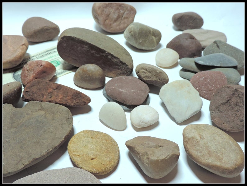 Pu&ograve; includere: Una collezione di pietre lisce e arrotondate in varie tonalit&agrave; di marrone, grigio e rosso. Le pietre sono disposte su una superficie bianca.