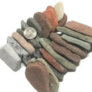 May include: A collection of smooth, flat stones in various shades of red, brown, gray, and green. The stones are stacked in a pile, with a US quarter coin for scale.