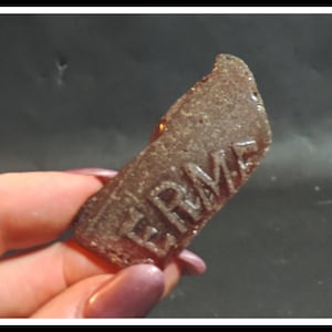 May include: A close-up of a brown glass shard with the word "ERMA" embossed on it.