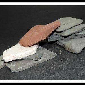 May include: A collection of flat, irregularly shaped stones in various colors including a smooth reddish-brown piece, a white stone, and several layered gray slate-like rocks stacked together to form a small, textured, and natural arrangement.