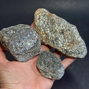 May include: Three rough, natural stones with a shimmering, metallic appearance. The stones vary in size and shape, with a mix of gray and gold tones. The largest stone is approximately 5 inches long. The stones are held in a hand against a dark background.