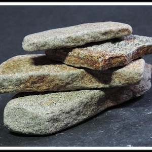 May include: A stack of four weathered, flat stones. The stones are primarily gray with patches of brown and reddish-brown. The surfaces are textured and irregular, suggesting natural erosion. The stones are arranged in a slightly offset, layered configuration.