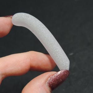 May include: A curved, frosted glass object, held between fingers. The piece is a light gray color and has a textured surface. The background is a dark, solid color.