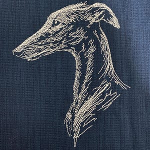 Puede incluir: Una tela azul marino oscuro que presenta un contorno bordado detallado de color claro de un galgo de perfil. El bordado utiliza puntadas finas y texturizadas para definir la forma de la cabeza, el hocico largo y el cuello esbelto del perro.