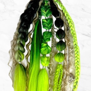 Könnte beinhalten: Ein Set aus grünen, schwarzen und blonden Haarverlängerungen mit Perlen und Charms. Die Verlängerungen sind mit einem schwarzen Gummiband zusammengebunden.