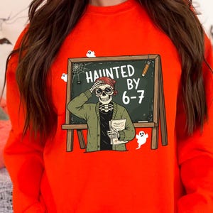 May include: An orange Halloween-themed sweatshirt featuring a skeleton teacher in front of a chalkboard that reads "Haunted by 6-7". The skeleton wears a green jacket and a red bandana. White ghost and spiderweb graphics are also present.