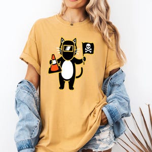 May include: Mustard yellow t-shirt featuring a cartoon cat wearing a welding mask and holding a traffic cone and a skull and crossbones flag. The cat is black with a white belly. The shirt has rolled-up sleeves.