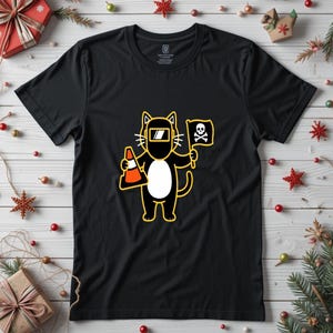 May include: Black t-shirt featuring a cartoon cat wearing a welding mask and holding a traffic cone and a flag with a skull and crossbones. The cat is black with a white belly and yellow outline. The flag is yellow with a black skull and crossbones.