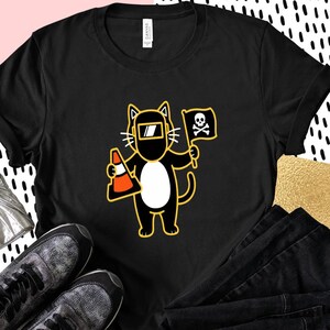 May include: Black t-shirt featuring a cartoon cat wearing a welding mask and holding a traffic cone and a flag with a skull and crossbones. The cat has a white belly and yellow outline.