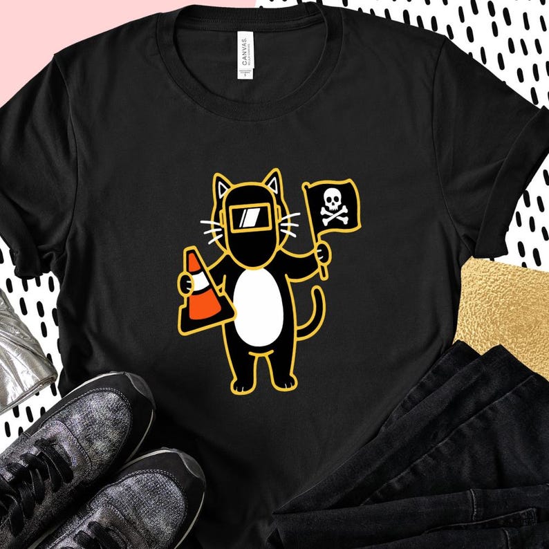 May include: Black t-shirt featuring a cartoon cat wearing a welding mask and holding a traffic cone and a flag with a skull and crossbones. The cat has a white belly and yellow outline.