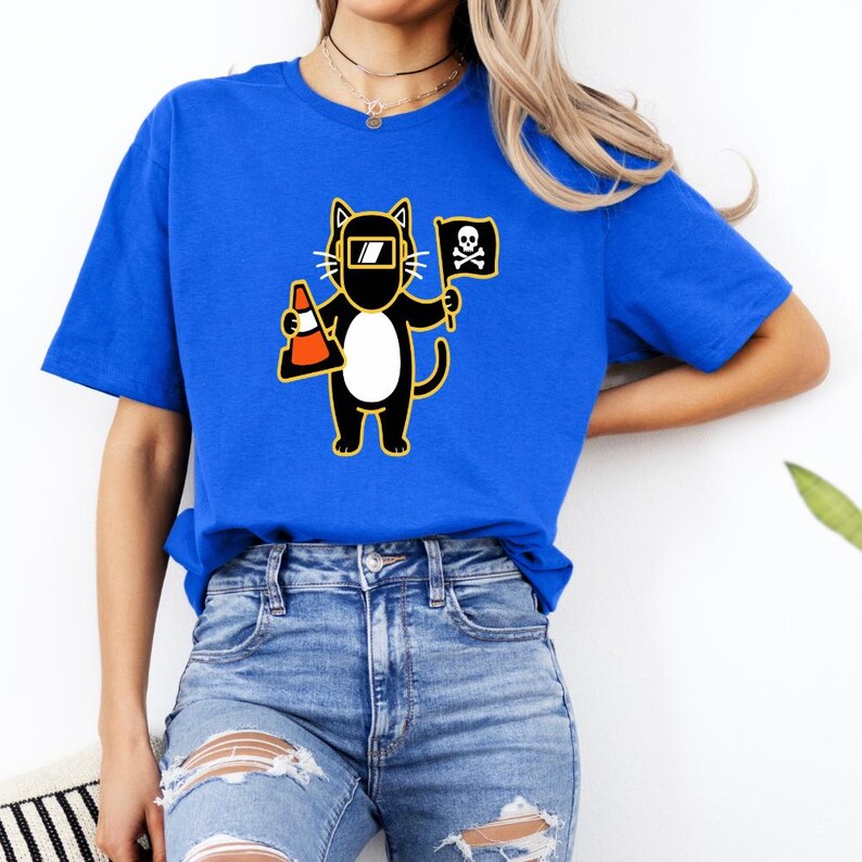 May include: Royal blue t-shirt featuring a cartoon cat wearing a welding mask and holding a traffic cone and a pirate flag with a skull and crossbones. The cat is black with a white belly and yellow outline.