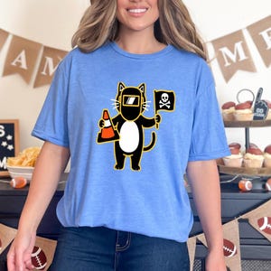 May include: Light blue t-shirt featuring a cartoon cat wearing a welding mask and holding a traffic cone and a pirate flag. The cat is black with a white belly and yellow accents. The flag has a skull and crossbones.