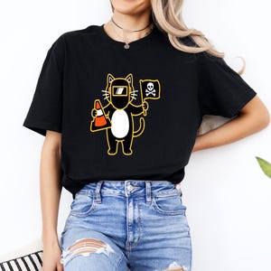 May include: Black t-shirt featuring a cartoon cat wearing a welding mask and holding a traffic cone and a pirate flag with a skull and crossbones. The cat has a white belly and yellow outline.