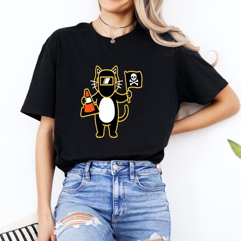 May include: Black t-shirt featuring a cartoon cat wearing a welding mask and holding a traffic cone and a pirate flag with a skull and crossbones. The cat has a white belly and yellow outline.