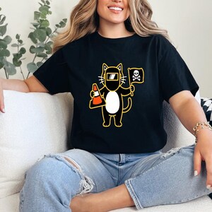 May include: Black t-shirt featuring a cartoon cat wearing a welding mask and holding a traffic cone and a flag with a skull and crossbones. The cat is outlined in yellow with a white belly. The shirt is a casual wear item.