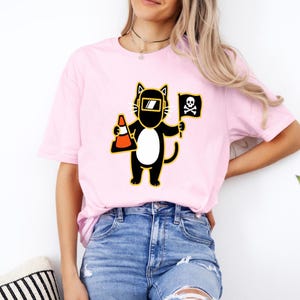 May include: A light pink t-shirt featuring a cartoon cat wearing a welding mask and holding a traffic cone and a pirate flag. The cat is black and white with yellow accents. The flag has a skull and crossbones.