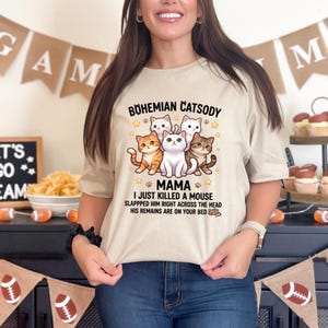 Może przedstawiać: Beżowa koszulka z grafiką przedstawiającą pięć kreskówkowych kotów i napisem "Bohemian Catsody Mama". Poniżej tekst: "I just killed a mouse slapped him right across the head his remains are on your bed".