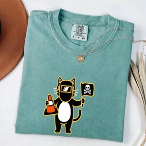May include: A teal Comfort Colors t-shirt featuring a cartoon cat wearing a welding mask and holding a traffic cone and a pirate flag with a skull and crossbones. A silver necklace with a pendant is on the shirt.