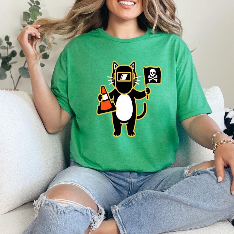 May include: Green t-shirt featuring a cartoon cat wearing a welding mask and holding a traffic cone and a pirate flag. The cat is black with a white belly and yellow accents. The flag has a skull and crossbones.