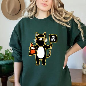 May include: Dark green sweatshirt featuring a cartoon cat wearing a welding mask and holding a traffic cone and a flag with a skull and crossbones. The cat is black with a white belly and yellow accents. The sweatshirt is a solid color.