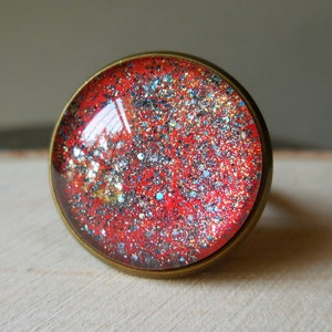 Op de afbeelding: Een rode ring met een glinsterend, galaxy-achtig ontwerp. De ring is gemaakt van metaal en heeft een ronde, platte bovenkant.