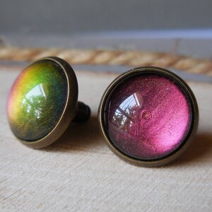 May include: A pair of round, iridescent glass earrings with a bronze metal setting. One earring has a green and yellow iridescent finish, while the other has a pink and purple iridescent finish.