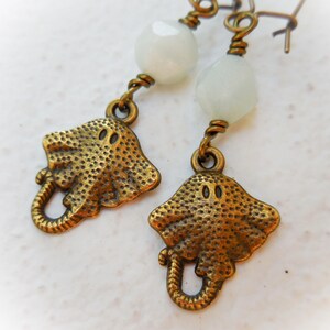May include: A pair of earrings featuring a bronze stingray charm and a white stone bead. The earrings are made with wire and have a dangling design.