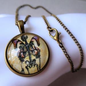 Könnte beinhalten: Eine Halskette aus Bronze mit einem runden Anhänger, der ein Skelett mit Schmetterlingsflügeln zeigt. Der Anhänger hat eine Glaskuppel und eine Vintage-Ästhetik.