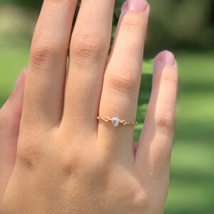 May include: A delicate gold ring with a small, round, white gemstone is worn on a finger. The ring features a thin chain design. The background is a blurred green, suggesting an outdoor setting.