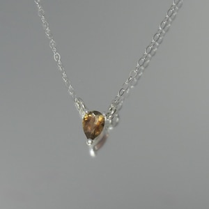 Könnte beinhalten: Eine zarte Silberkette mit einem tropfenförmigen, bernsteinfarbenen Anhänger. Der Anhänger hängt an einer feinen Kette, wodurch ein minimalistisches und elegantes Design entsteht. Der Edelstein hat einen warmen, goldenen Farbton.