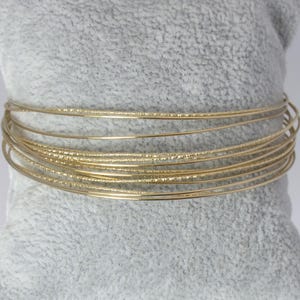 May include: A set of gold-colored bangles, stacked on a gray velvet pillow. The bangles have a thin, delicate design, with some featuring a textured surface. The jewelry is displayed against a neutral background, highlighting the gold color.