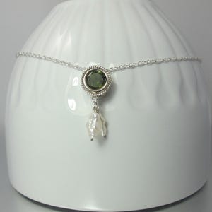 May include: A silver chain necklace featuring a circular pendant with a faceted olive green gemstone set in a twisted metal border. A small, irregular white baroque pearl hangs from the bottom of the circular setting, creating a delicate drop design.