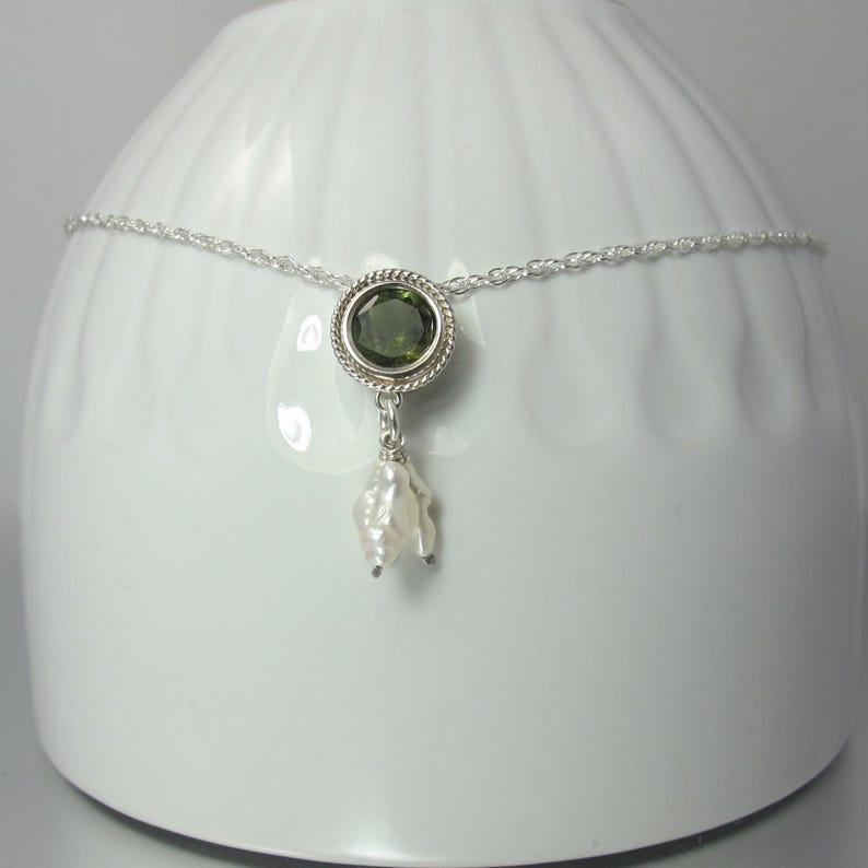 May include: A silver chain necklace featuring a circular pendant with a faceted olive green gemstone set in a twisted metal border. A small, irregular white baroque pearl hangs from the bottom of the circular setting, creating a delicate drop design.