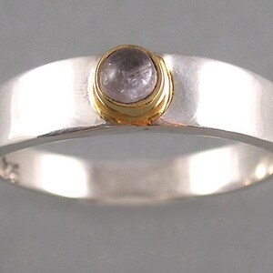 May include: A silver ring with a round, clear gemstone set in a gold bezel. The ring is simple and elegant.