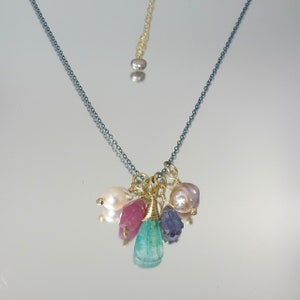 May include: A delicate gold chain necklace with a cluster of charms, including a large green gemstone, pink and white pearls, and a small purple stone.