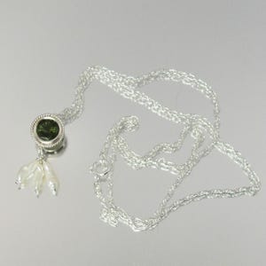 May include: A silver chain necklace featuring a circular pendant with a dark green faceted stone set in a twisted metal border. Three small, elongated white pearls hang from the bottom of the pendant, creating a delicate, dangling accent.