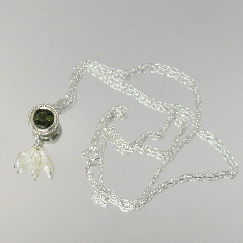 May include: A silver chain necklace featuring a circular pendant with a dark green faceted stone set in a twisted metal border. Three small, elongated white pearls hang from the bottom of the pendant, creating a delicate, dangling accent.