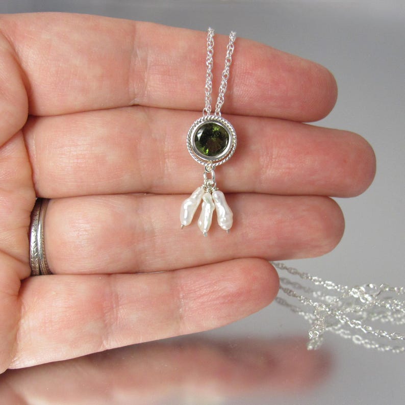 May include: A silver necklace featuring a circular pendant with a faceted olive green gemstone set in a twisted metal border. Three small, irregular white pearls hang from the base of the circular setting, suspended by a delicate silver chain.