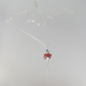 May include: A silver chain necklace featuring a pendant composed of a small blue bead, a flat silver disc, a faceted red stone, and a long, thin, pointed silver spike. The necklace is displayed against a plain, light-colored background.