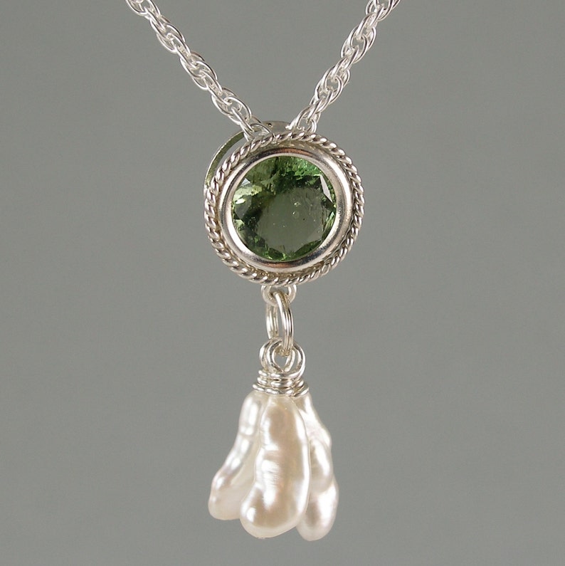 May include: A silver necklace featuring a round, faceted green gemstone set in a twisted metal frame, with a cluster of three elongated, irregular white pearls suspended from the bottom of the setting on a silver chain.